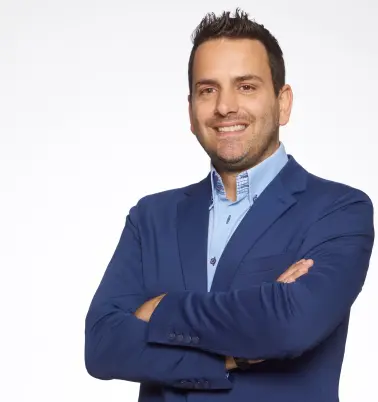 Ein Mann mit dunklen Haaren verschränkt die Arme vor der Brust. Er trägt ein dunkelblaues Jackett über einem hellblauen Hemd. Er lächelt.