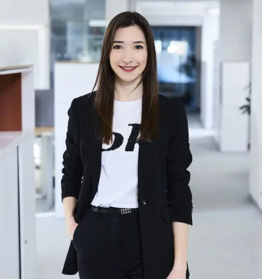 Eine Frau mit dunklen Haaren steht in einem Büroflur. Sie trägt einen schwarzen Blazer und hat eine Hand in der Hosentasche. Sie lächelt.