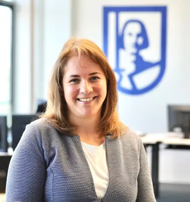 Frau mit braunen Haaren lächelt in die Kamera. Im Hintergrund ist ein Büro zu sehen mit einem großen ROLAND Assistance Logo.