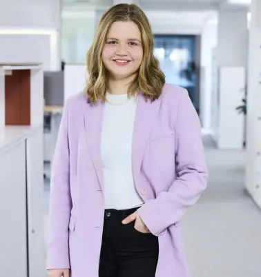 Eine Frau mit blonden Haaren steht in einem Büroflur. Sie trägt einen lilafarbenen Blazer und hat eine Hand in der Hosentasche. Sie lächelt.