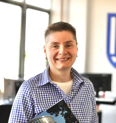 Frau mit kurzen Haaren steht in einem Büroraum. Sie schaut nach vorne und lächelt.