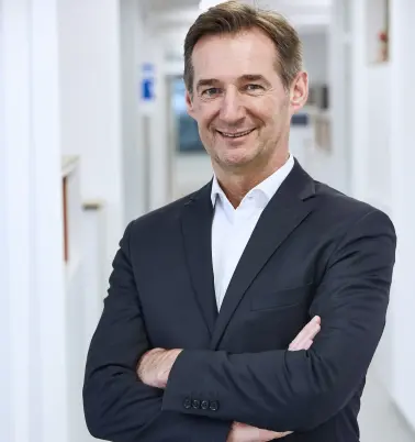 Ein Mann mit dunklen Haaren verschränkt die Arme vor der Brust. Er steht in einem Büroflur. Er trägt ein dunkles Jackett über einem weißem Hemd. Er lächelt.