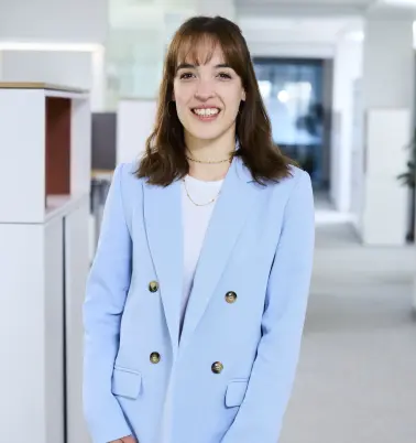 Eine Frau mit schulterlangen braunen Haaren steht in einem Büroflur. Sie trägt einen hellblauen Blazer. Sie lächelt.