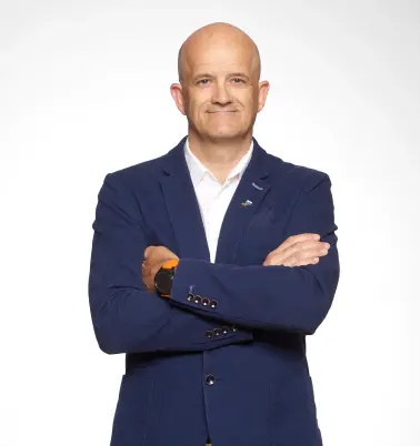 Ein Mann mit Glatze verschränkt die Arme vor der Brust. Er trägt ein dunkelblaues Jackett über einem weißen Hemd. Er lächelt.