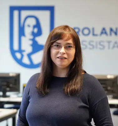 Frau mit braunen Haaren und Brille lächelt in die Kamera. Im Hintergrund ist ein Büro zu sehen mit einem großen ROLAND Assistance Logo.