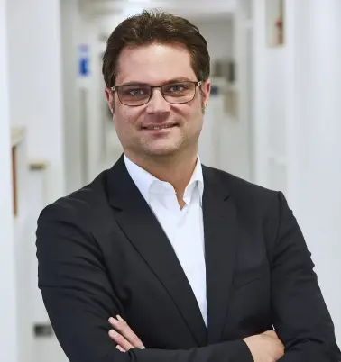 Ein Mann mit dunklen Haaren und Brille verschränkt die Arme vor der Brust. Er steht in einem Büroflur. Er trägt ein dunkles Jackett über einem weißem Hemd. Er lächelt.