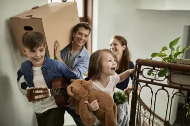 Famile zieht um und lacht. Sie steigen in einem Hausflur die Treppe herauf. Die zwei Kinder tragen Spielsachen, die Eltern tragen Kisten. 