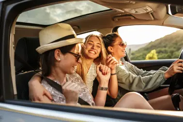 Drei junge Frauen sitzen gemeinsam im fahrenden Auto. Eine Frau die hinten sitzt umarmt die Beifahrerin. Alle drei lachen. Sie tragen sommerliche Kleidung und Sonnenbrillen. 