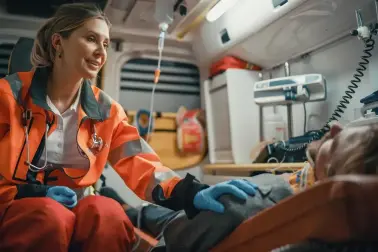 Junge Sanitäterin sitzt in einem Rettungswagen. Sie trägt Handschuhe und hat ein Stethoskop um den Hals. Eine Person liegt auf einer Liege neben ihr und trägt eine Halskrause. Sie legt dieser Person ihre Hand auf die Schulter.